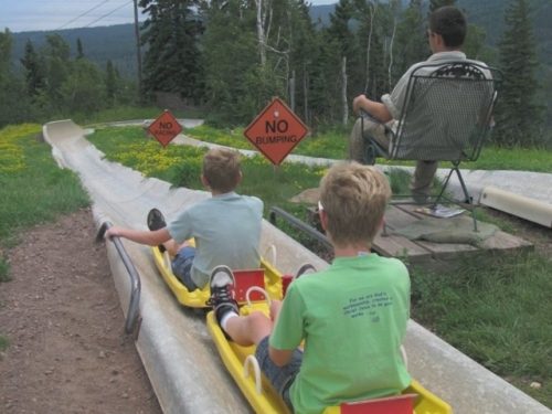 Two young riders on yellow sleds heading down neighboring alpine slide tracks past a trackside warning sign